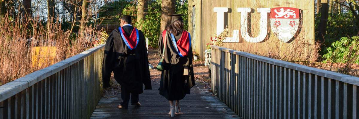 Two students wearing graduation gowns, walking on a wooden bridge with large LU letters behind