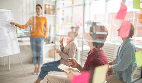 Three people attending a presentation photographed through a glass wall covered with post-it notes