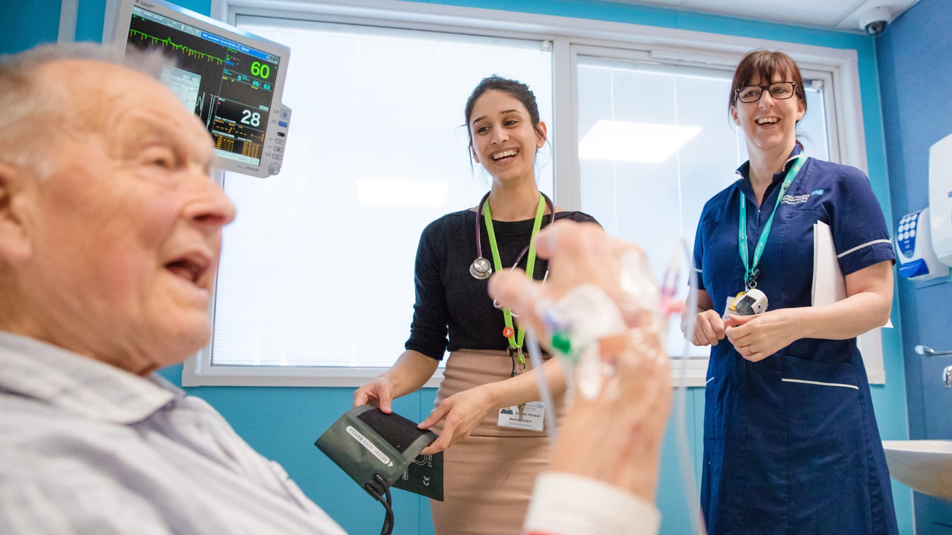 An old man in a hospital bed is watched by a laughing doctor and nurse.