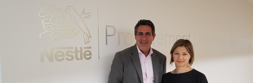 A man and a woman standing in front of a long beige wall with the Nestlé logo and text reading "[text obscured by peoples' heads] Delivering Value to our Bu..."