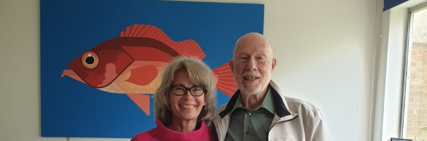 White woman in pink top, and older white man, smiling, standing in front of a stylized painting of a carp.