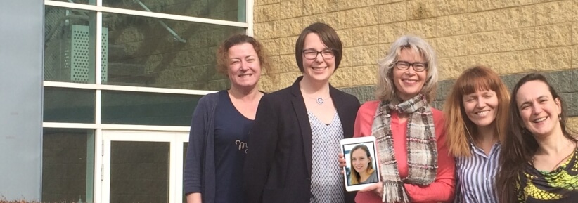 5 women standing together in front of a wall and a glass door, holding a picture of a 6th woman.