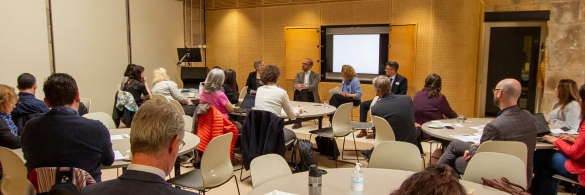 People sitting round tables in a meeting room, attention on four presenters at the front of the room