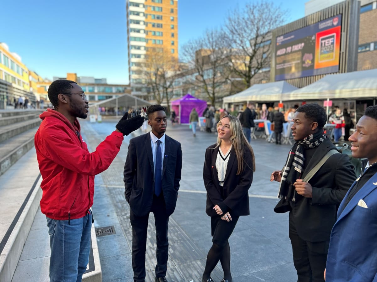 Two students sat at bench having chat, 2 more students sat behind on wall