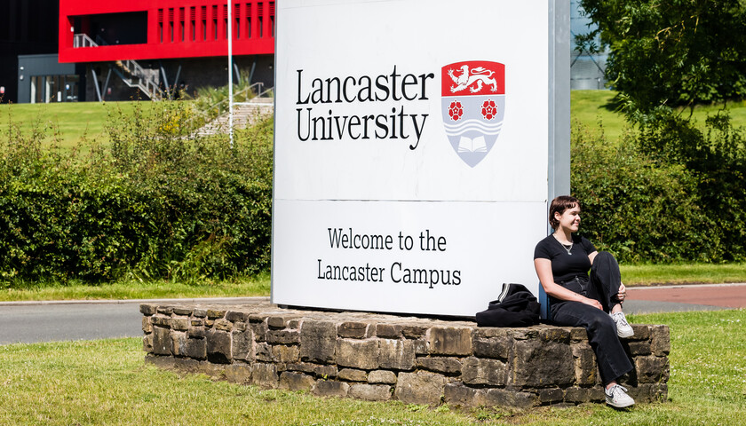 A students relaxes in the sun next to the main Lancaster University sign