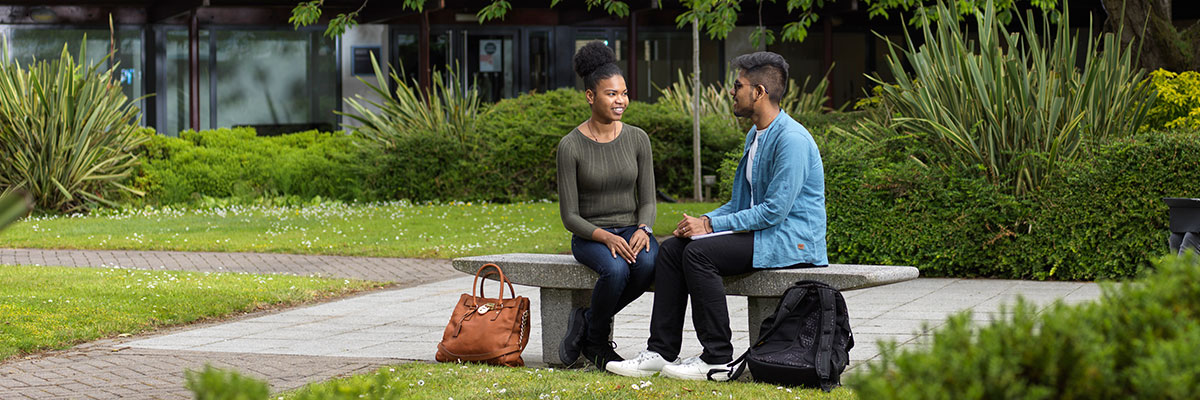 two students sit on a bench in a leafy university quad