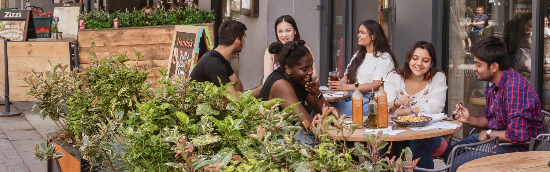 Students sitting at a restaurant table are surrounded by tables and foliage.