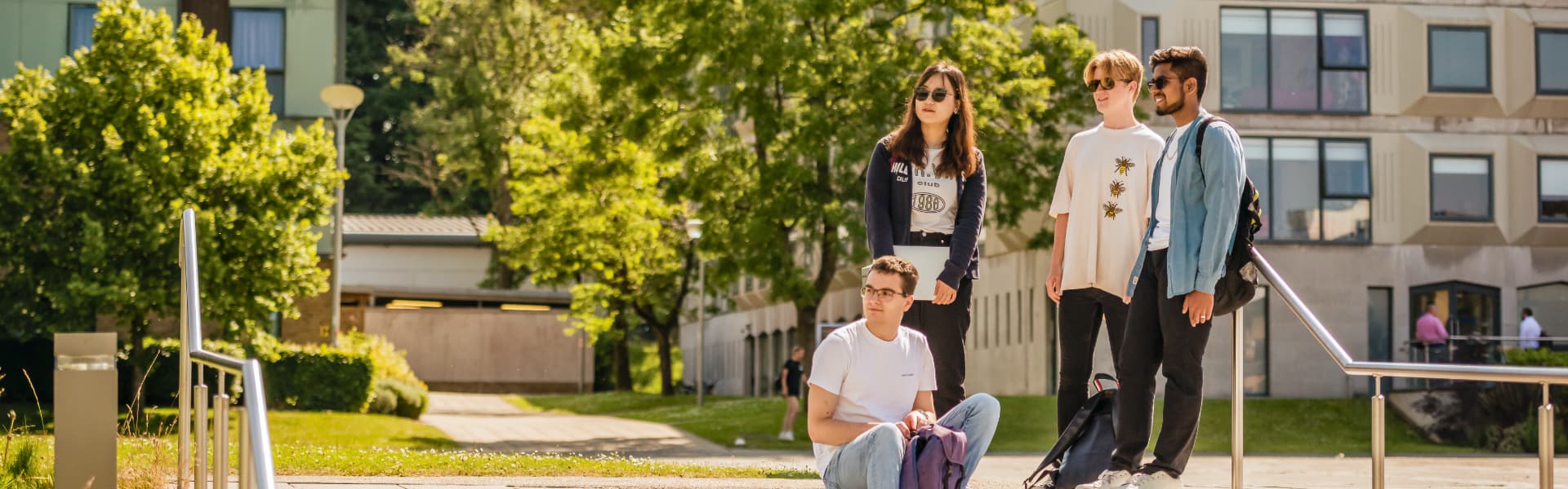 Four students stand and sit by the LICA building in the sunshine, wearing sunglasses.