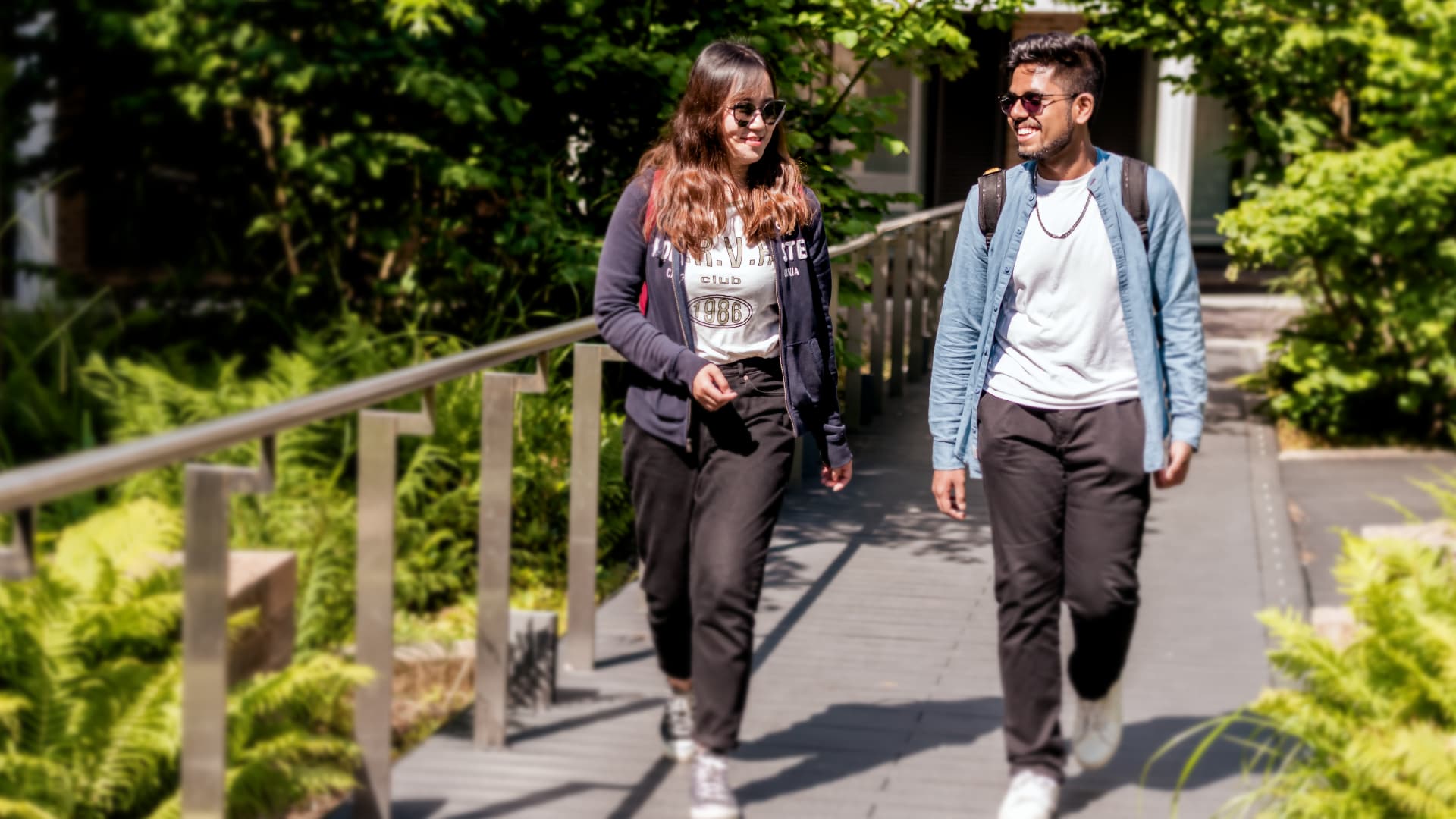 Two students walk through ferns in a sunny spot on campus