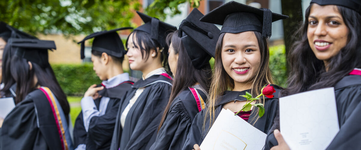 Students stand outside in mortar boards and gowns, clutching certificates.