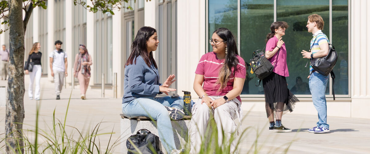 Students in groups stand outside the Management School building in the sunshine.