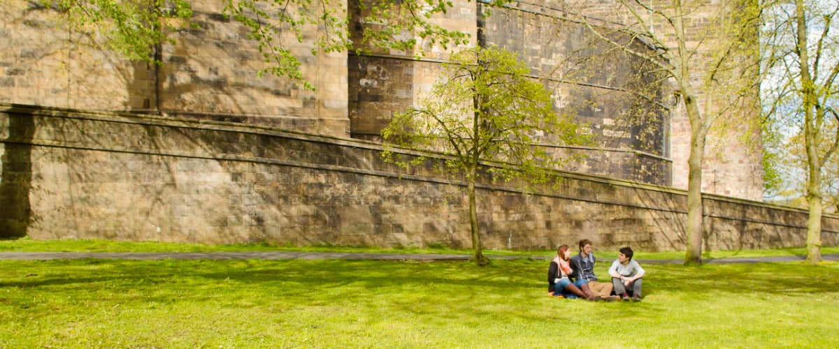Three students sit on the grass in front of the ancient stone walls of Lancaster Castle.