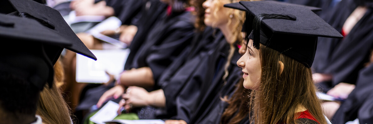 A row of students sits in a large hall in academic caps and gowns.