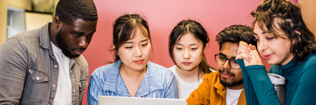 A group of five students stand around a laptop. They are smiling and laughing.