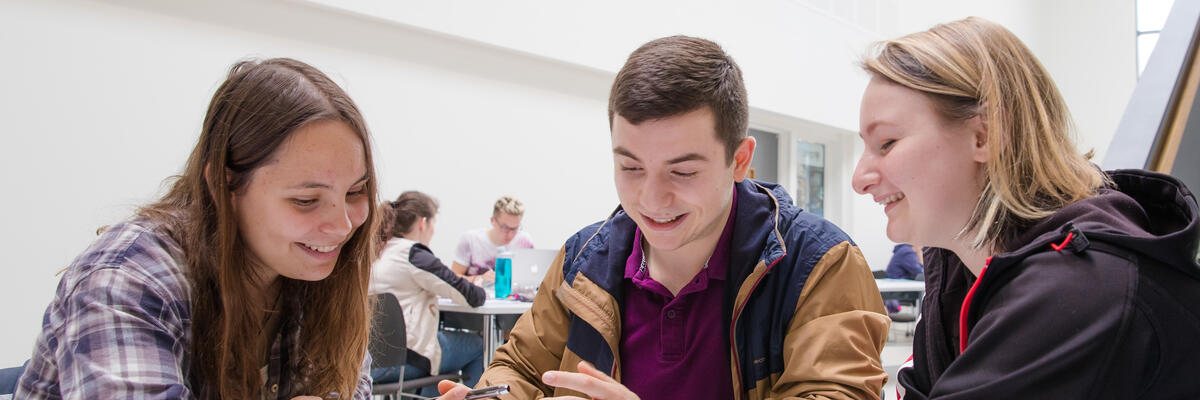 Three students laugh whilst working around a table.