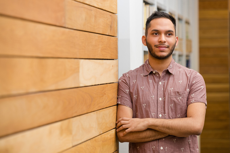 A male student leaning against a wall with arms folded facing the camera