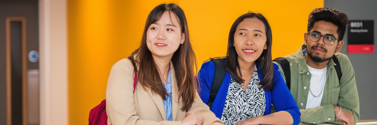 Three students lean over a balcony against a bright yellow background.