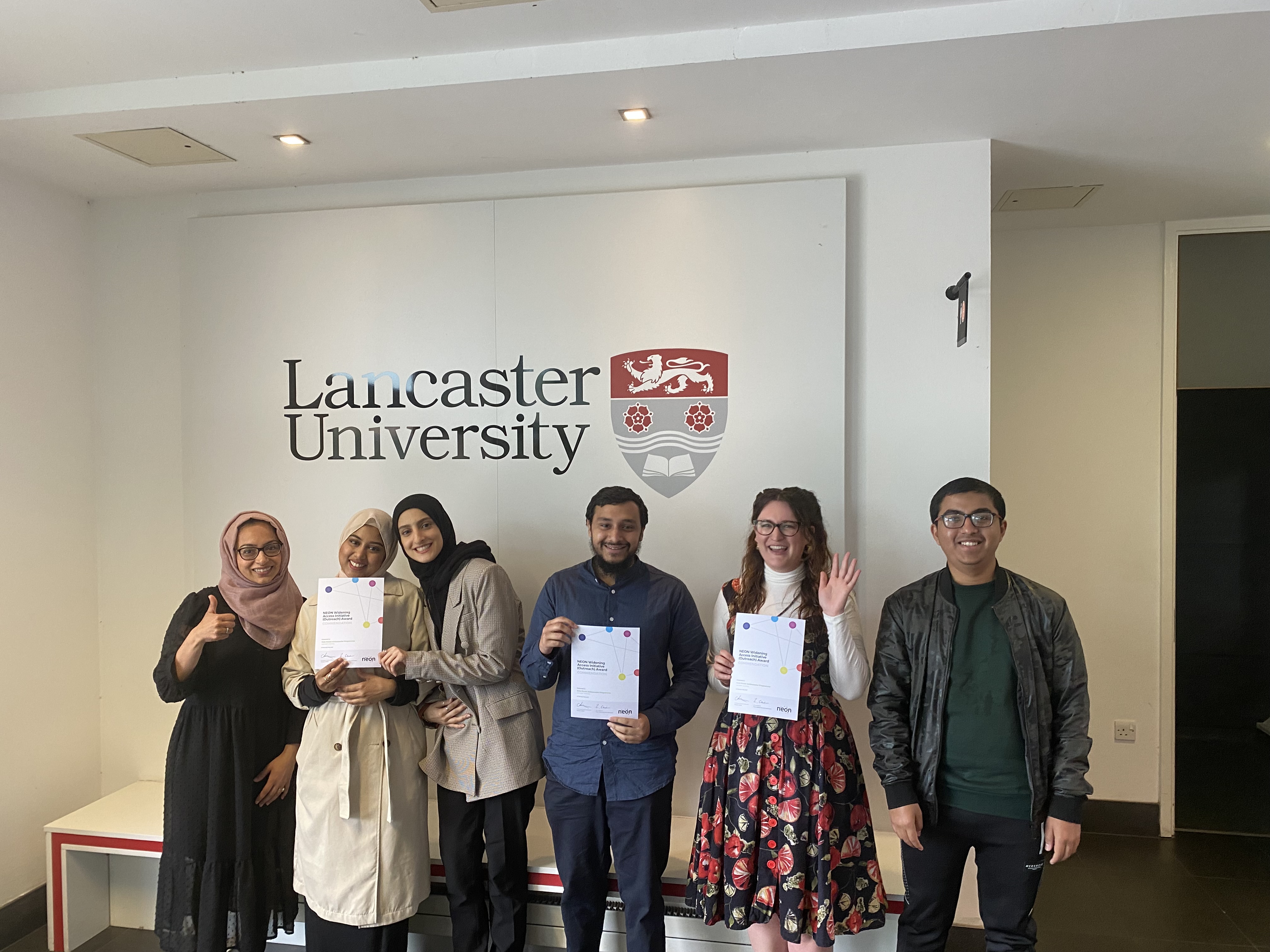 A group of students celebrate with certificates of achievement alongside staff. They are standing under a Lancaster University logo.